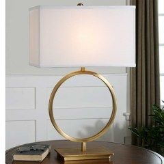 Gold ring table lamp with white rectangular shade on a wooden table near curtains.