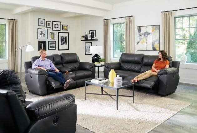 Living room with three black recliners, a coffee table, and two people seated near large windows.