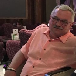 Man in a peach shirt and glasses smiling while seated in a patterned chair indoors
