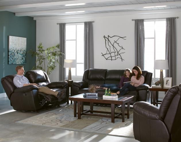 Living room with three people sitting on dark leather sofas around a coffee table near tall windows.