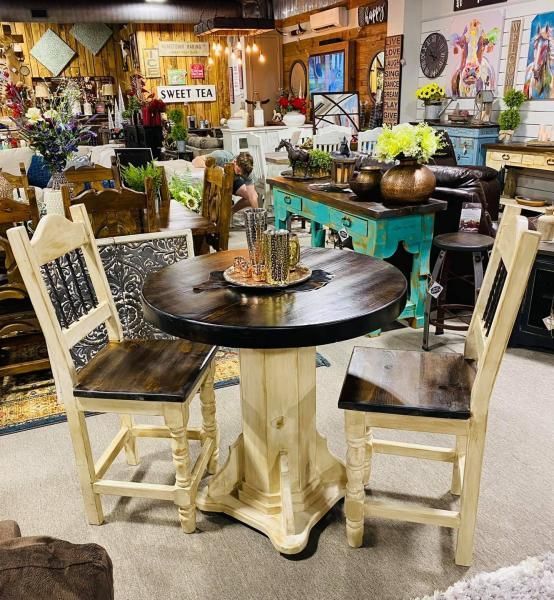 Rustic dining display with round black table, cream chairs, flowers, and vintage decor in a shop.