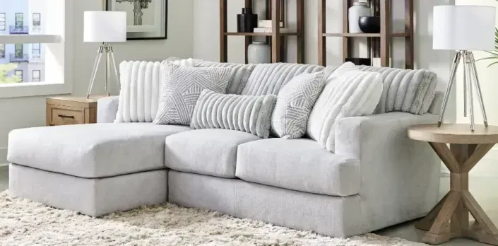 Light gray sectional sofa with white pillows in a bright living room, with lamps and shelving behind it
