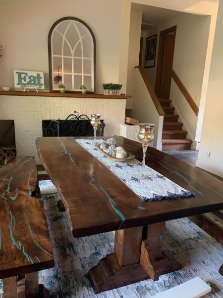 Rustic dining room with long wooden table, bench, staircase, and decorative shelf display.