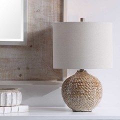 Textured table lamp with white shade on a white shelf beside stacked books and a framed wall print