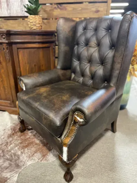 Black tufted leather wingback chair with gold nailhead trim in a room with a wooden cabinet and shag rug