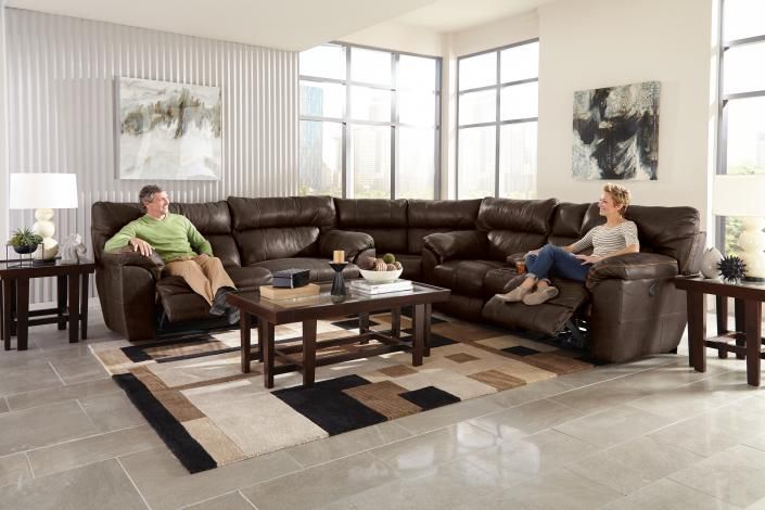 Two people sitting on a brown sectional in a bright living room with large windows and a coffee table
