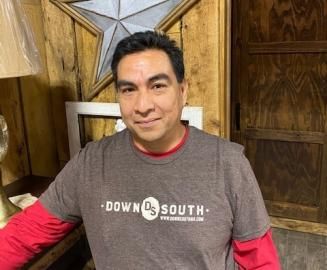 Man standing indoors in a brown Down South T-shirt, smiling near wood-paneled walls.