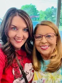 Two smiling women posing outdoors, one in a red shirt and one in a teal top and glasses.