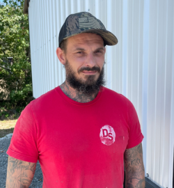Man in red T-shirt and cap standing outdoors beside a white wall, looking at the camera.