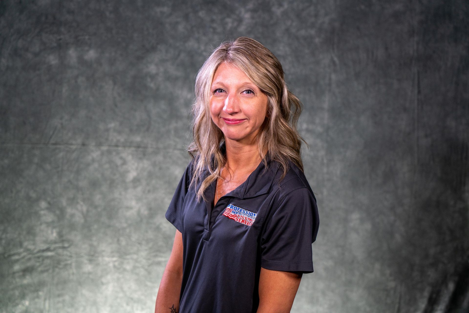 Woman in dark polo shirt with logo, smiling against a gray backdrop.