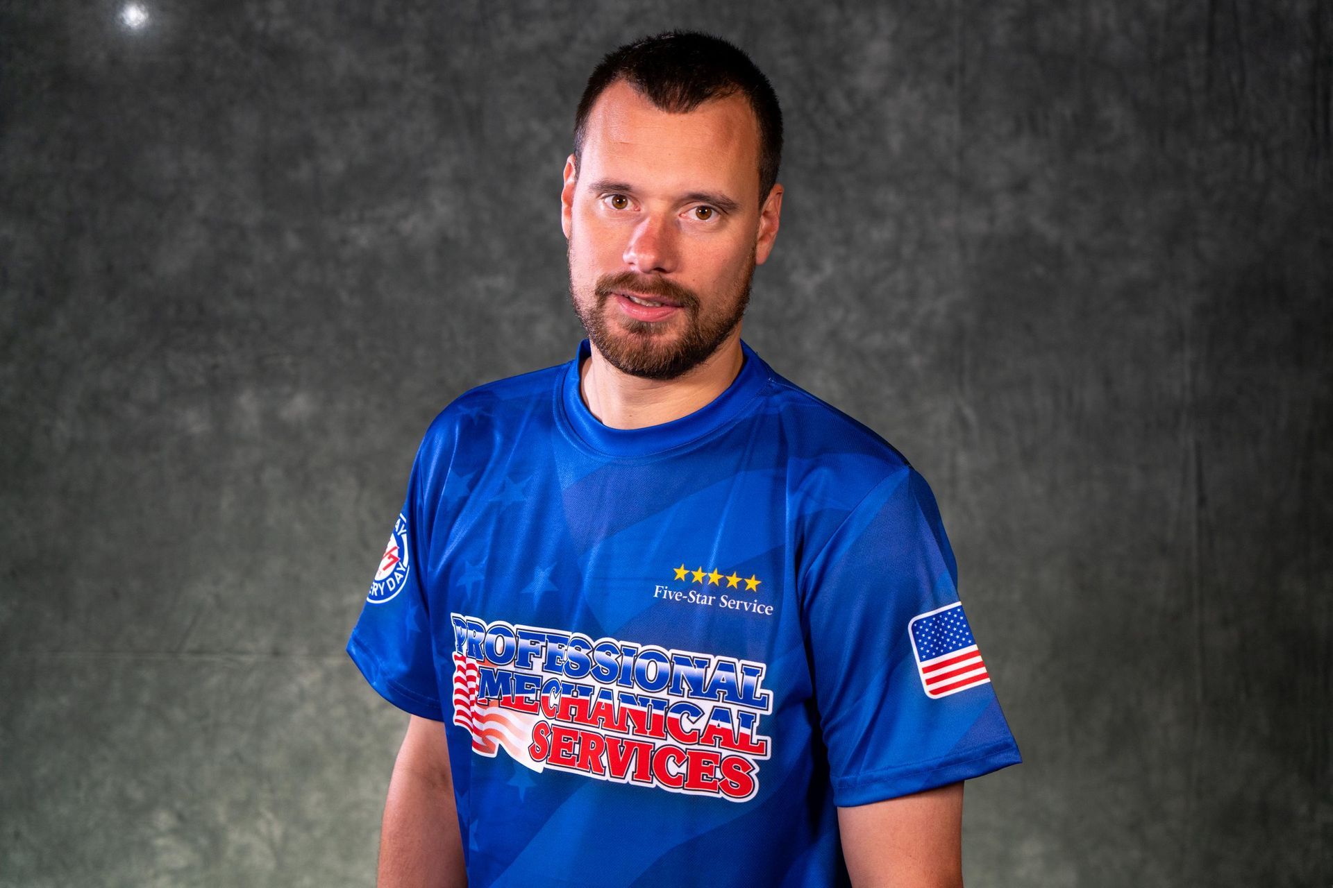 Man in blue shirt, American flag patch, logo on chest, grey background.