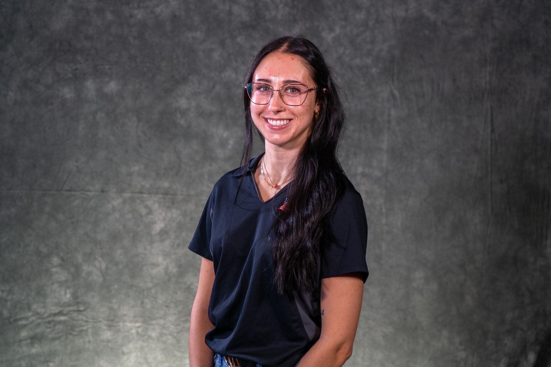 Woman in glasses smiles, wearing a black shirt, standing in front of a gray backdrop.