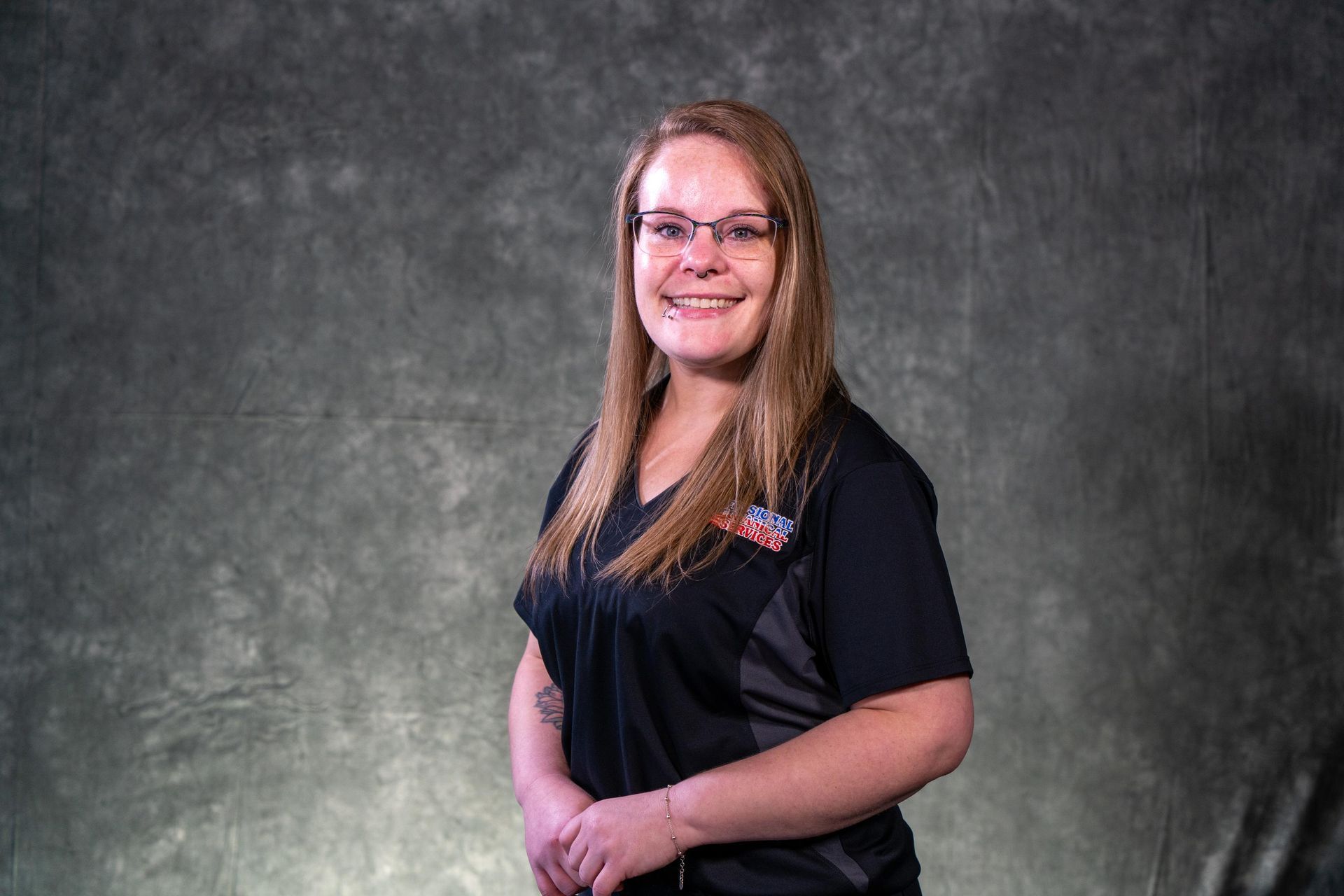 Woman with glasses, smiling, wearing a black shirt with grey accents, standing in front of a gray backdrop.