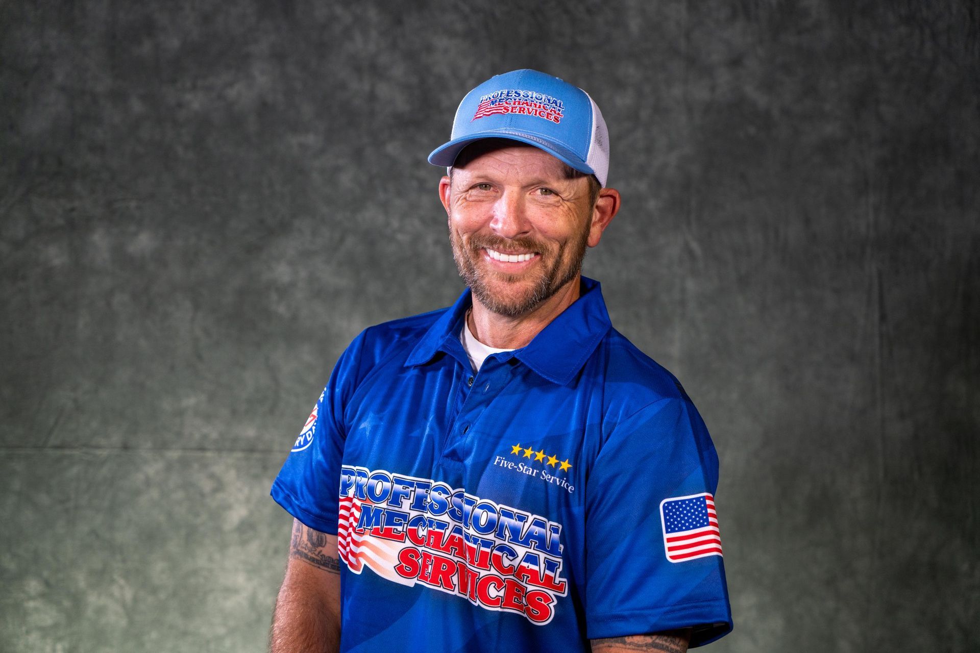 Man in blue shirt and cap smiles, with American flag on shirt sleeve, gray background.