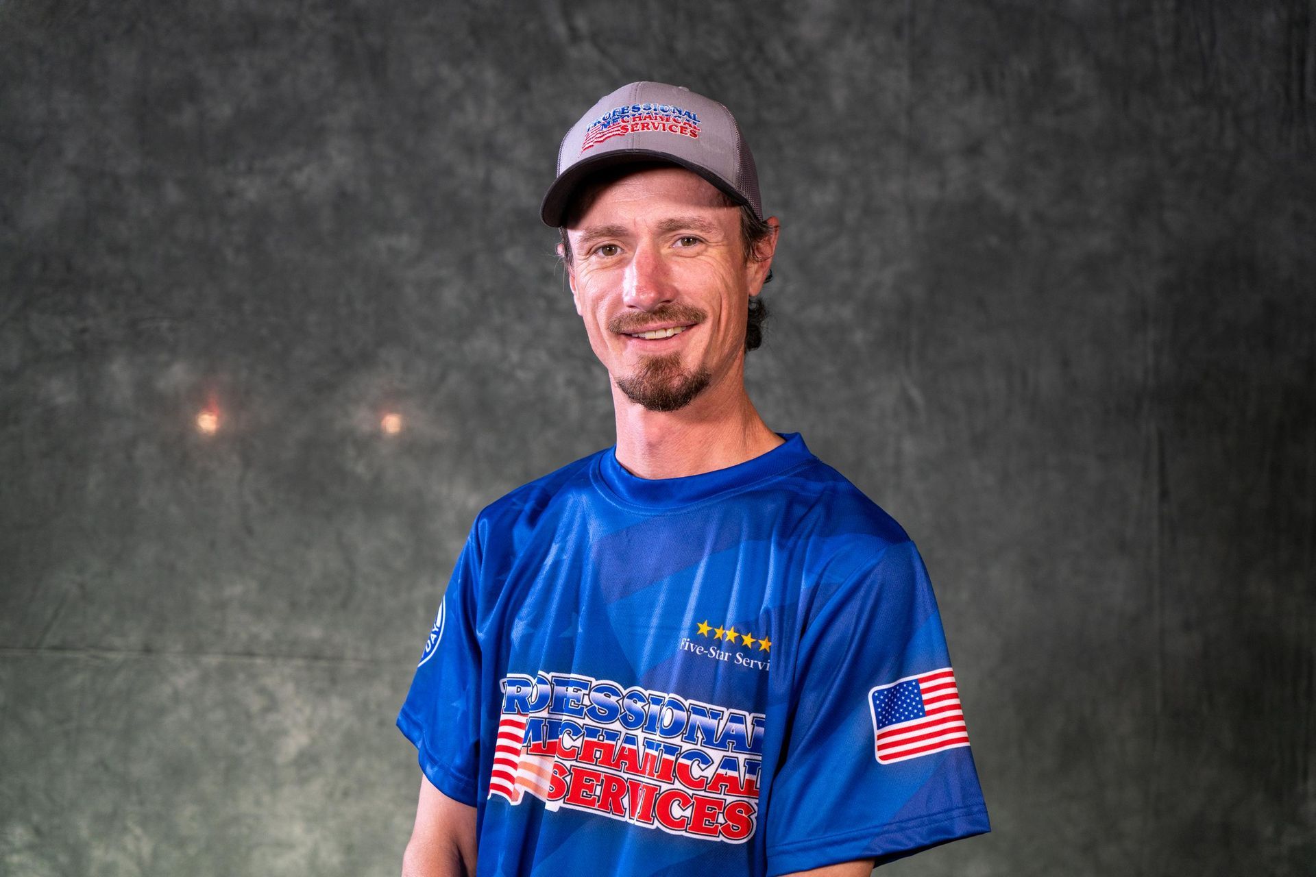 Man in blue shirt and cap smiles, set against a mottled gray background.