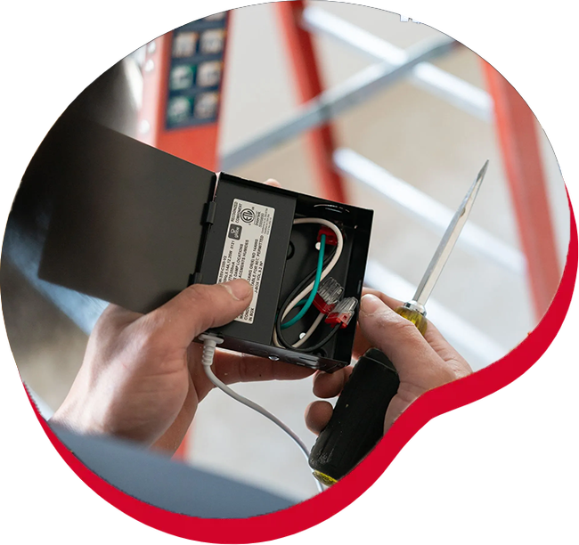 Person wiring electrical box with a screwdriver; red and white wires visible, ladder in background.