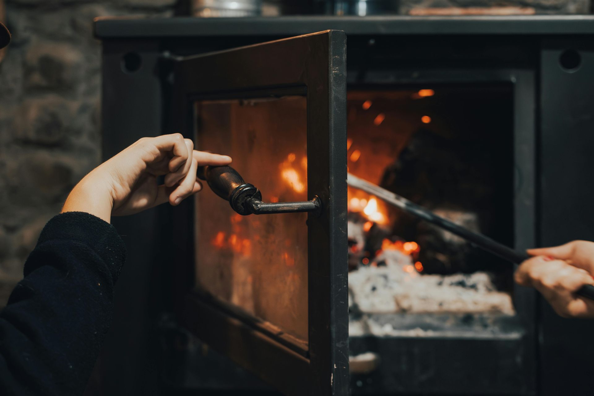 Person opening a wood-burning stove to tend to a fire inside, using a long tool.