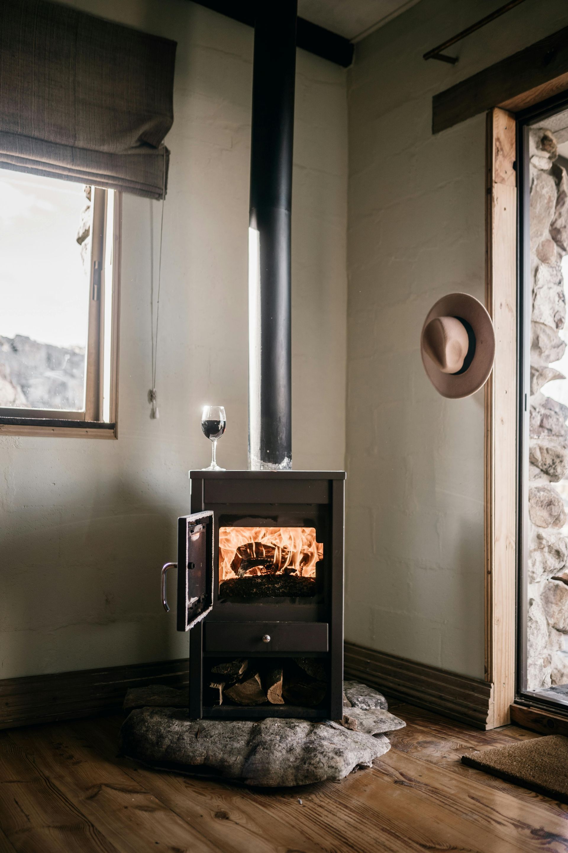 Wood-burning stove in a cabin with lit fire. A glass of wine on top. A hat hangs on wall.