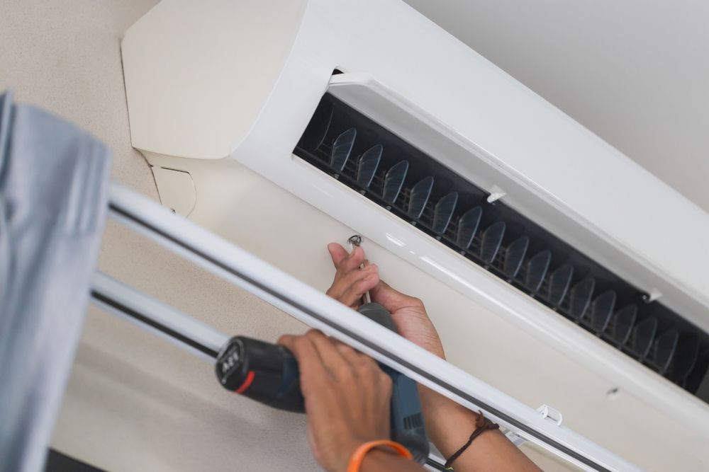 Person using a power drill to work on a white air conditioner unit mounted on a ceiling.