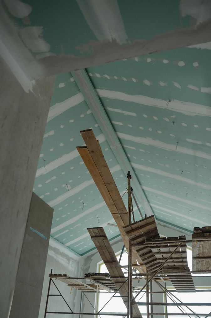 Worker installing drywall in a room with a ladder and exposed wiring.