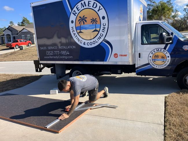 A technician kneels on a driveway, cutting material next to a Remedy Heating & Cooling truck.