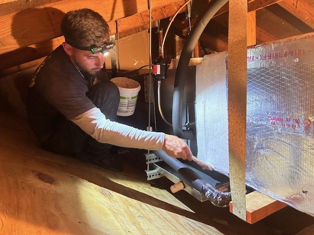 Man inspecting HVAC system in attic. Wearing headlamp. Holding a pipe.