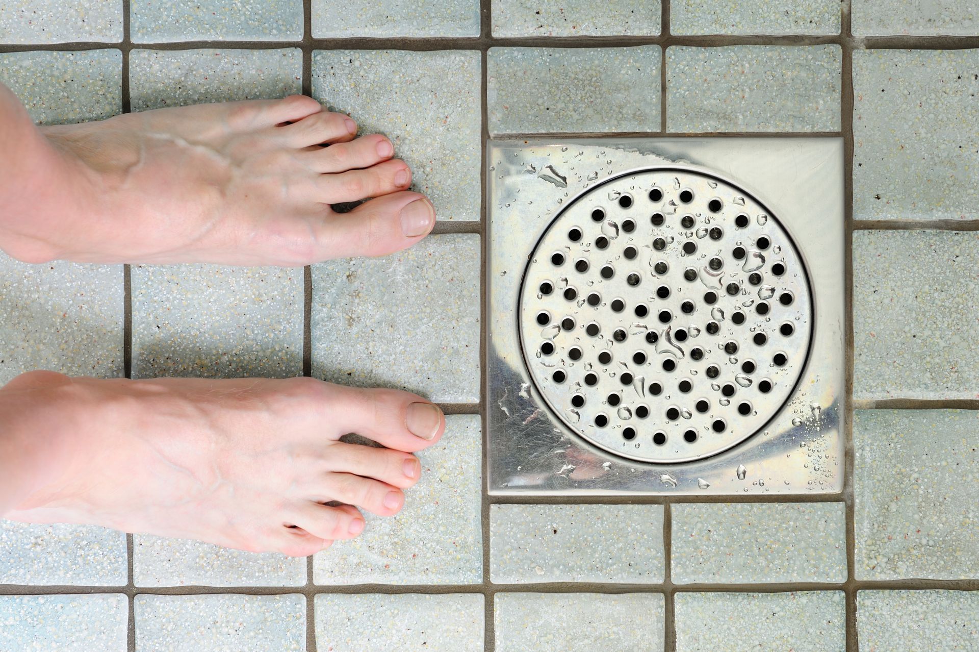 A person 's feet are standing next to a drain on a tiled floor. A person 's feet are standing next to a drain on a tiled floor.