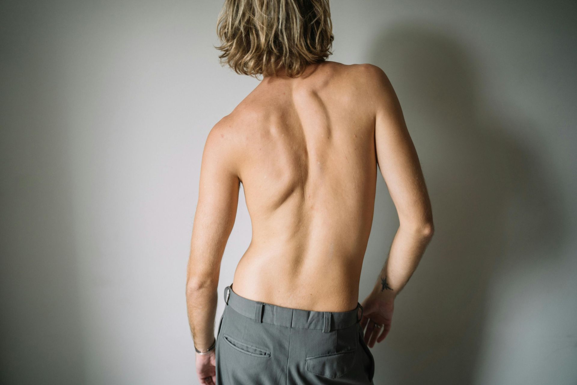 Back view of a person standing shirtless against a plain wall, wearing gray pants