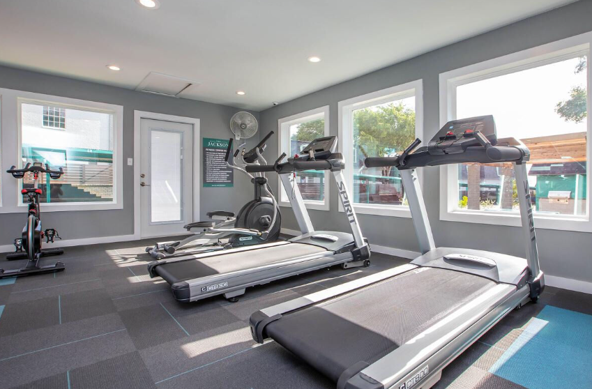Bright gym room with treadmills, elliptical machines, and large windows letting in natural light