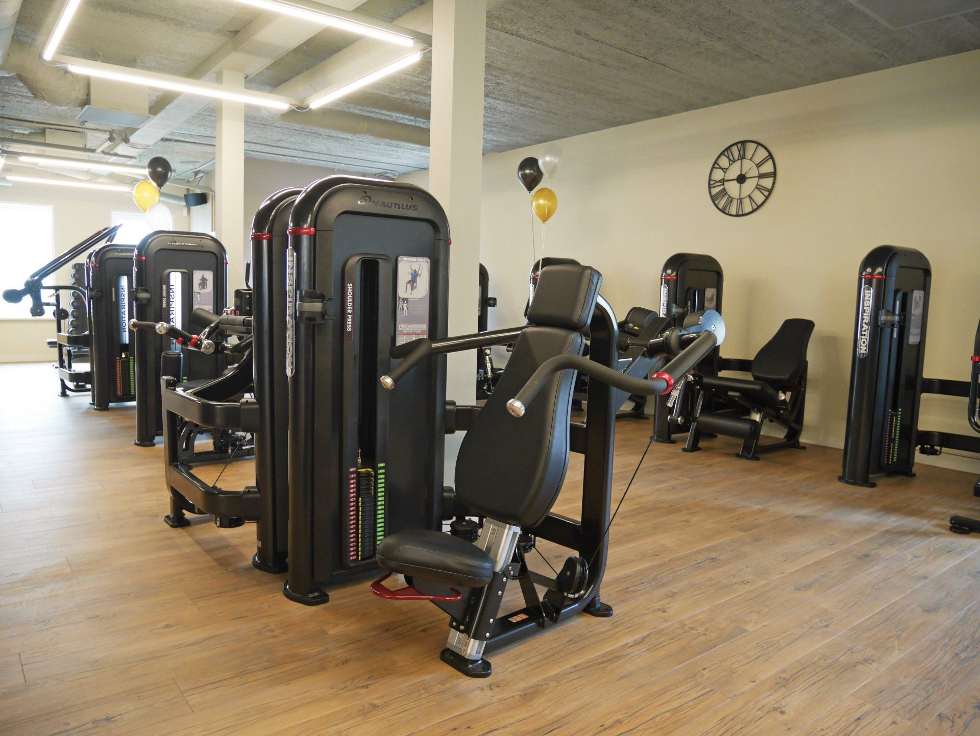 Sportschoolinterieur met fitnessapparaten, waaronder een zwarte shoulder press, op een lichte houten vloer.