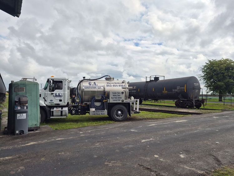 Utility trucks and a black tanker trailer parked on a paved lot under cloudy skies.