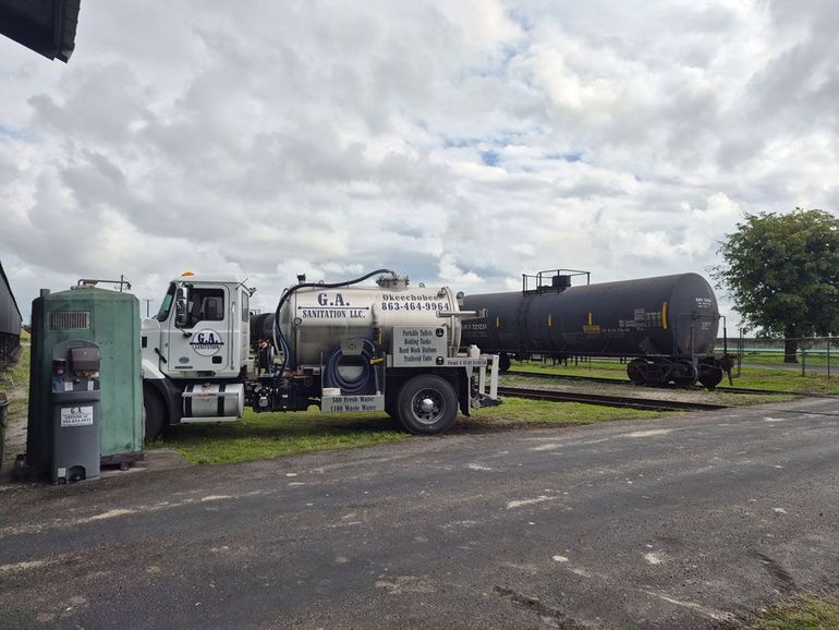 Utility trucks and a black tanker trailer parked on a paved lot under cloudy skies.