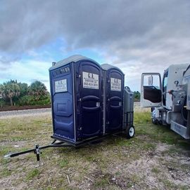 Two blue portable toilets on a trailer beside a utility truck in a grassy field