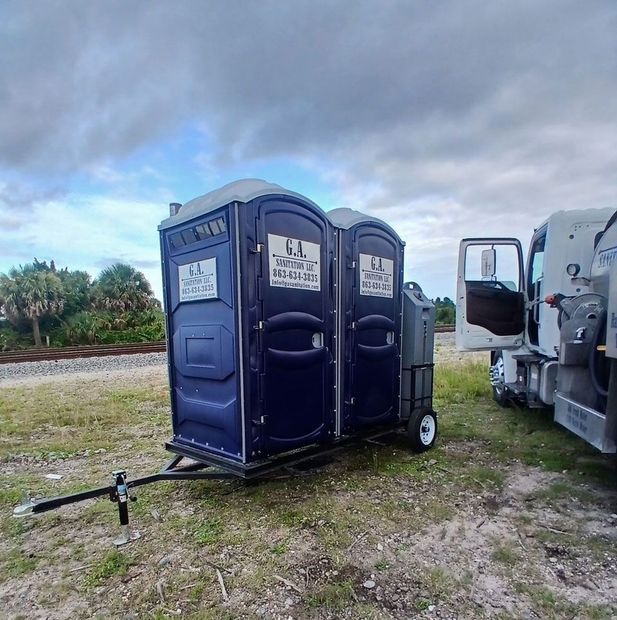 Two blue portable toilets on a trailer beside a utility truck in a grassy field