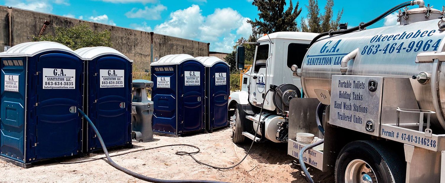 Blue portable toilets lined up beside a white utility truck in a dusty outdoor lot.