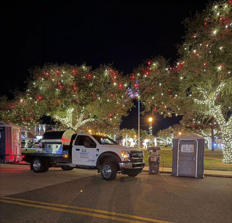 Garland-lit trees at night with a utility truck parked beside portable toilets on a street