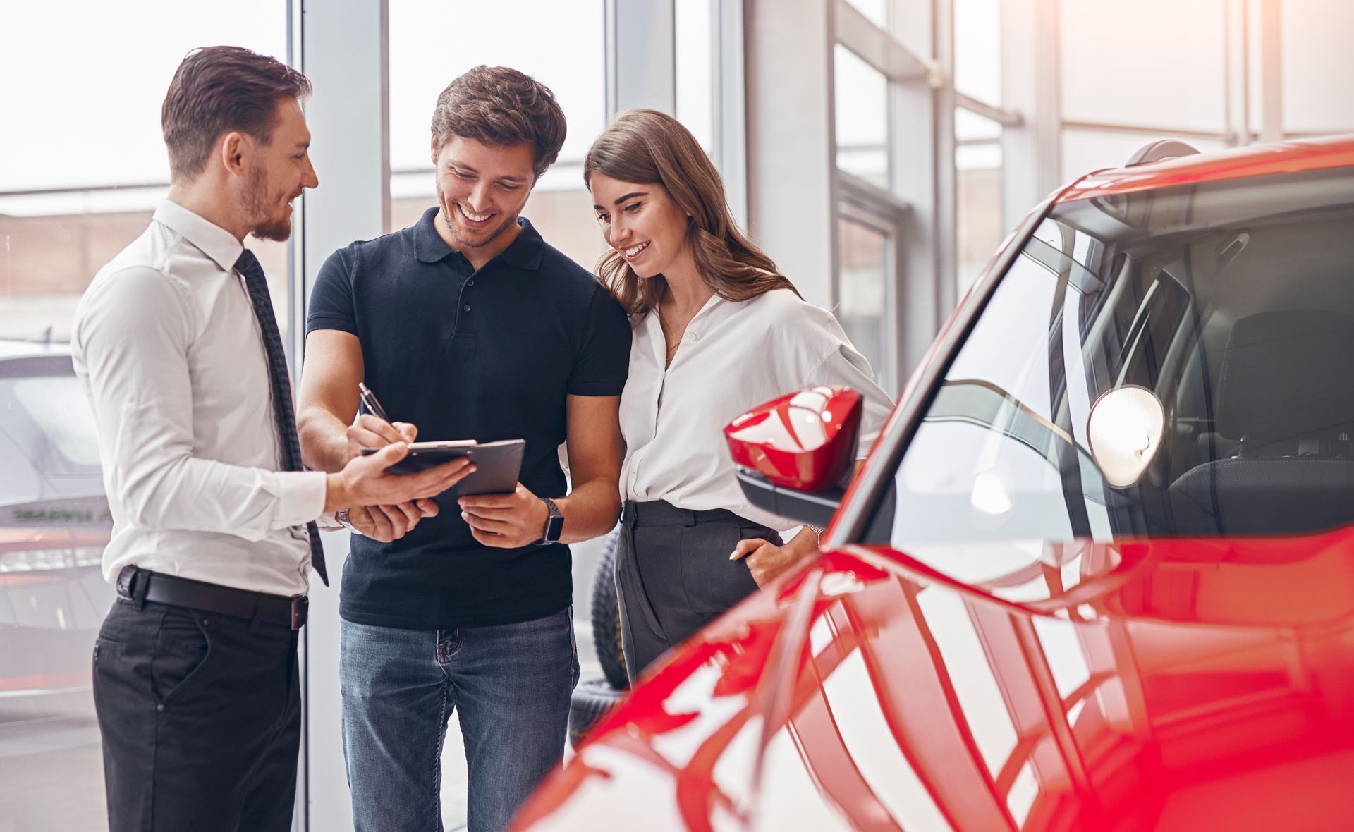 Three people reviewing paperwork beside a red car in a bright showroom