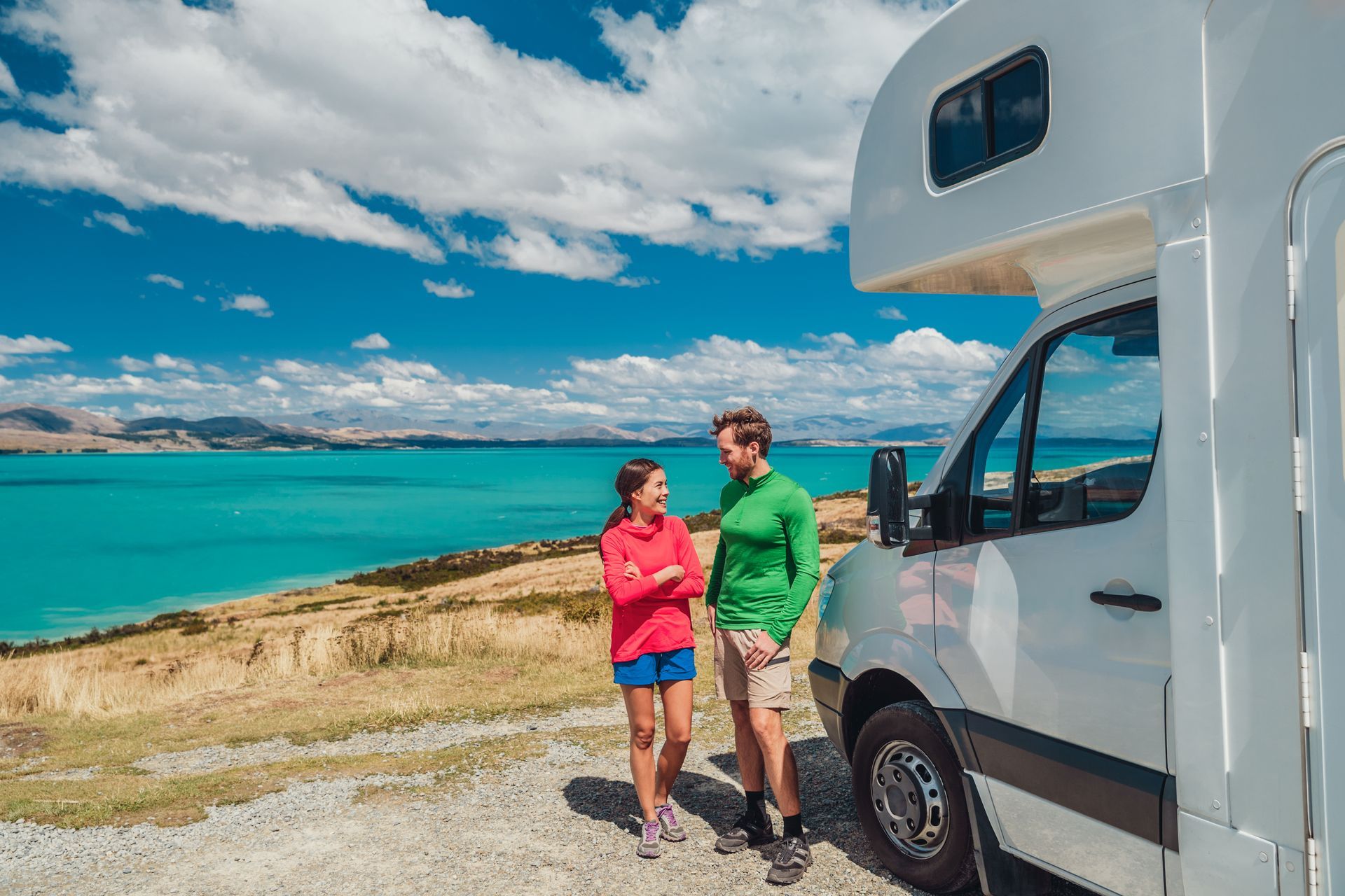 Two people stand beside a white camper van overlooking a turquoise sea under a partly cloudy sky.