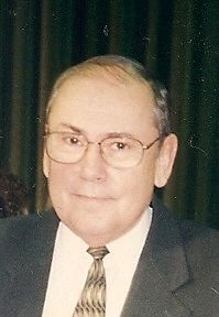 A man wearing glasses and a suit and tie is standing in front of a green curtain.