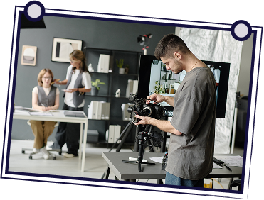 A person operating a video camera on a tripod, filming two people in a studio setting.
