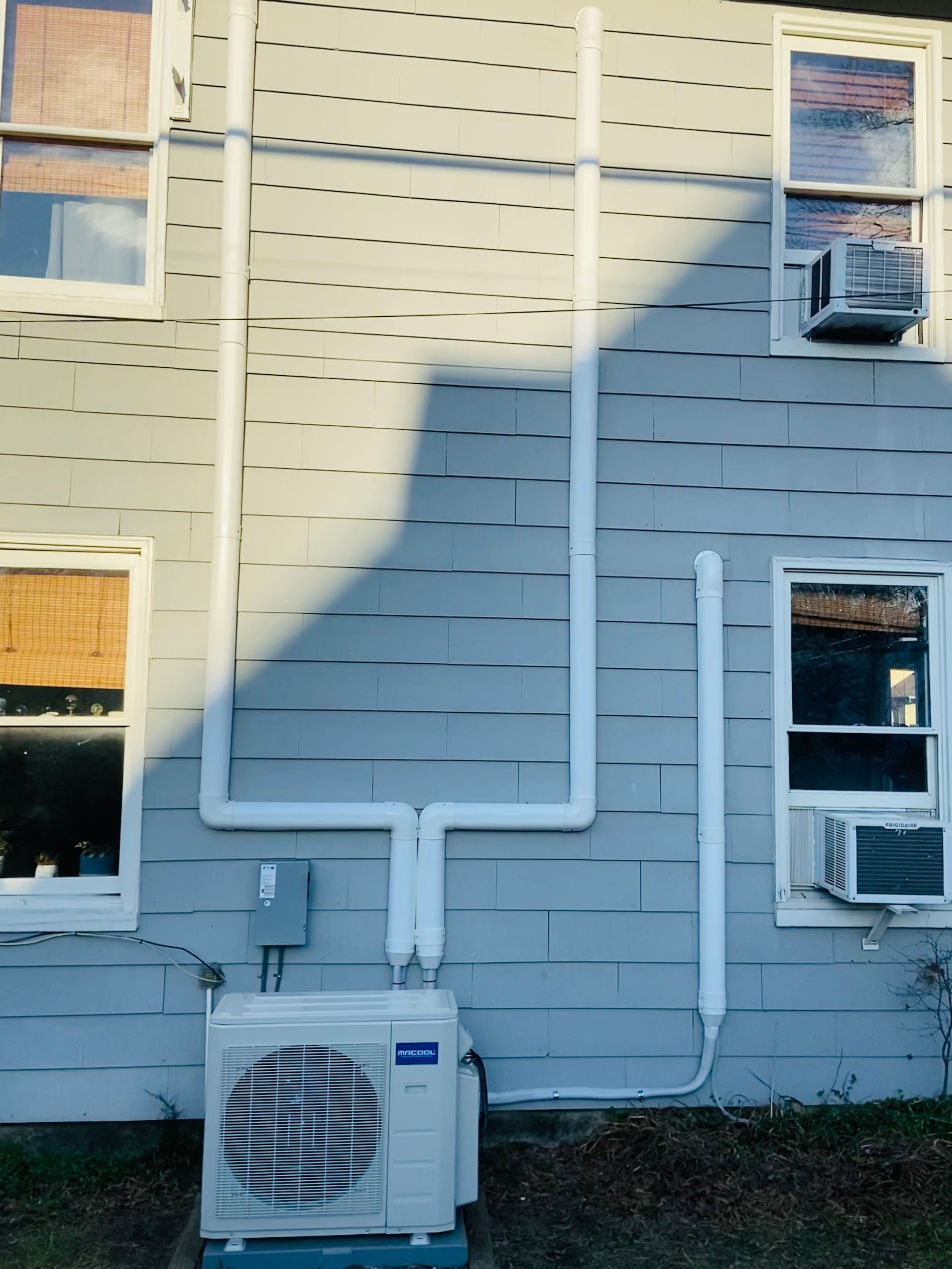 Building exterior with white utility pipes, an air conditioner unit, and a window air conditioner in sunlight