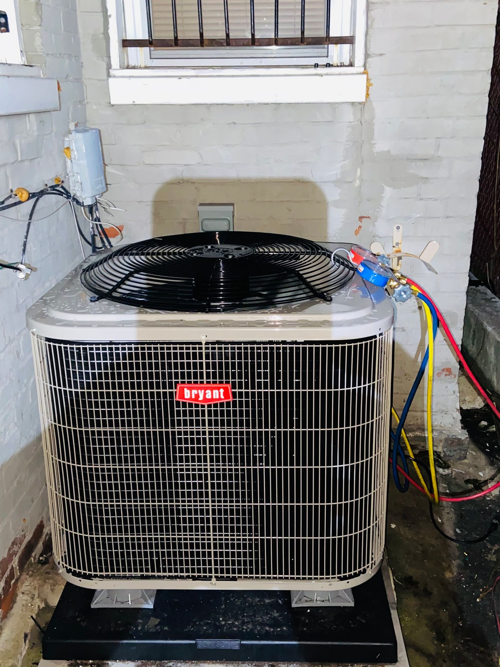 Outdoor air conditioning unit with exposed colorful wires beside a white brick wall
