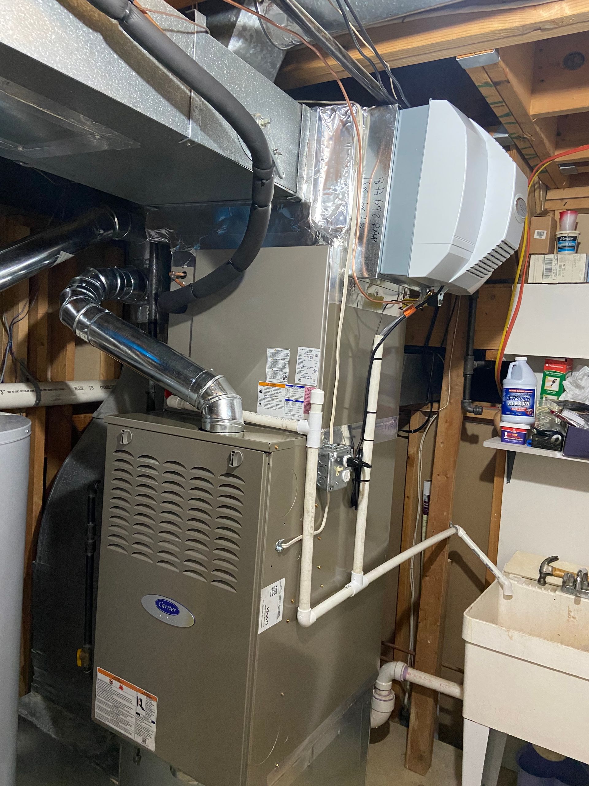 Furnace and ductwork in a cramped utility closet with white PVC piping and storage shelves.