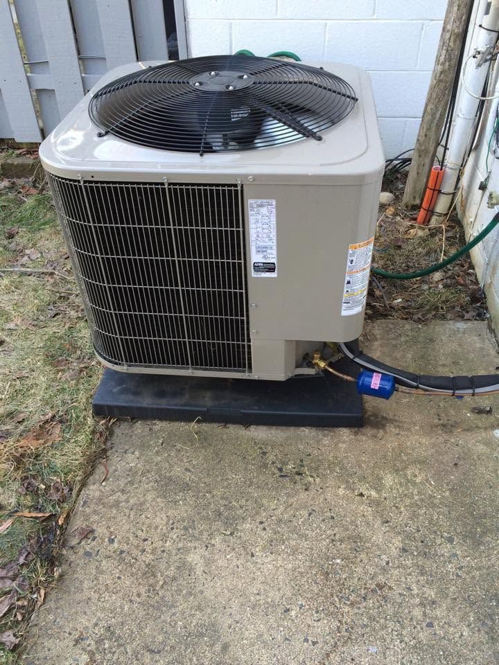 Outdoor central air conditioning unit on a concrete pad beside a house, with visible hoses and wiring.