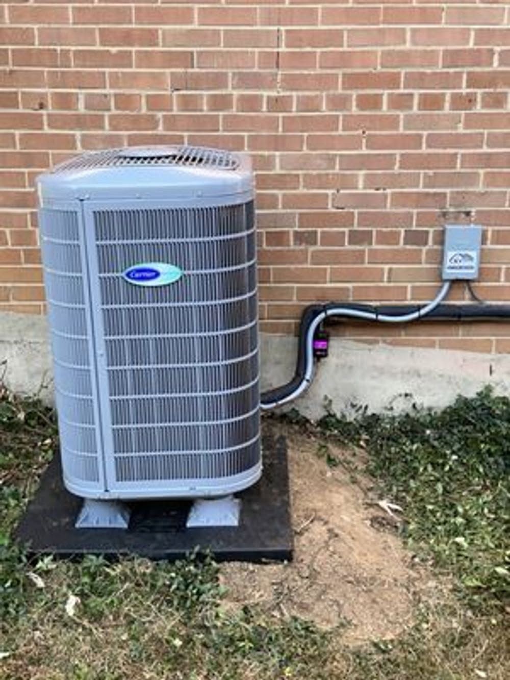 Outdoor air conditioner unit beside a brick wall, with conduit and electrical box on a small pad.