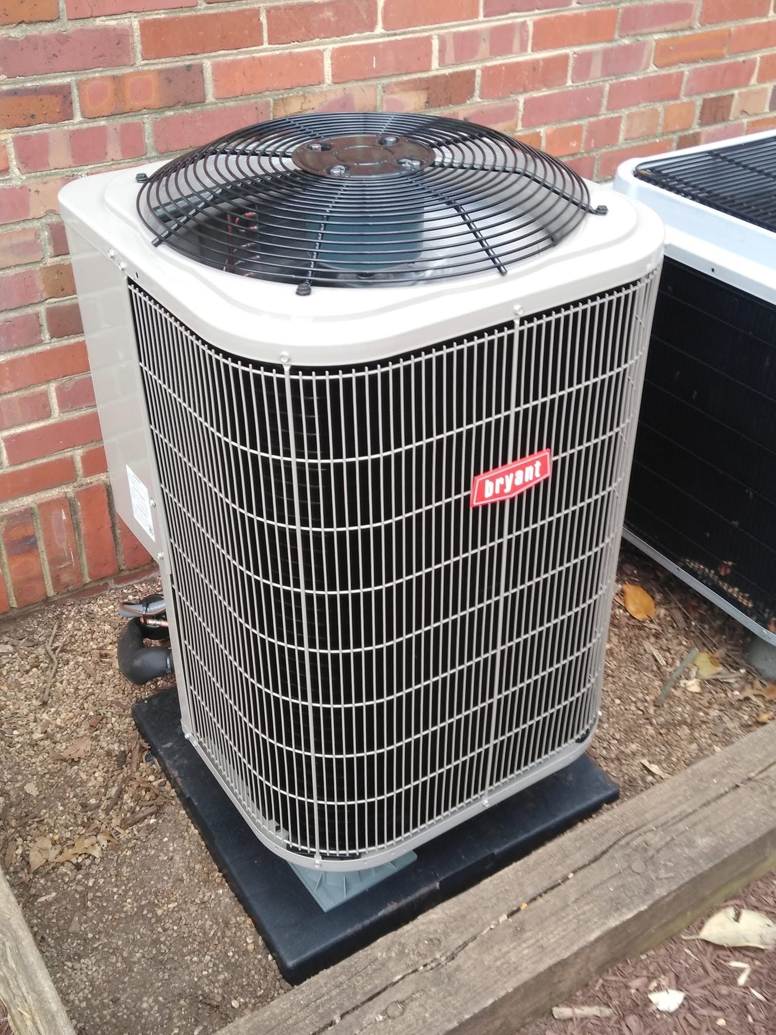 Outdoor air conditioning unit beside a brick wall, with a black fan grille and white metal sides.