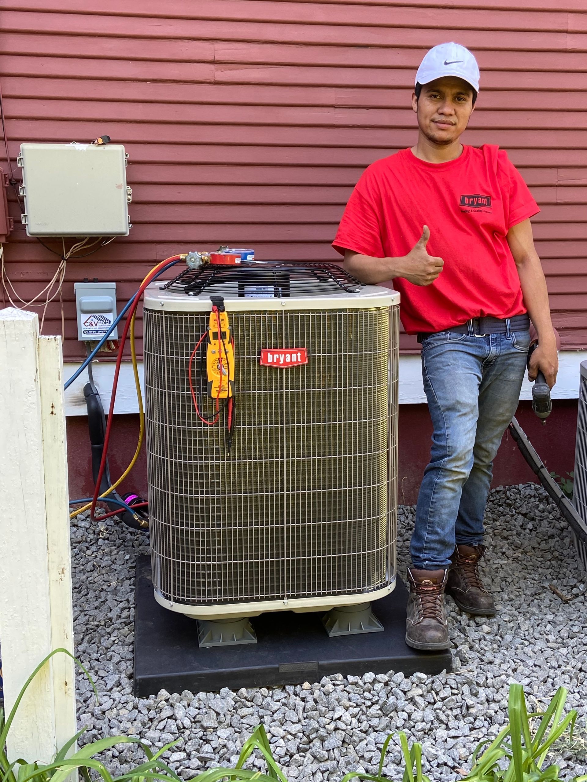 Man standing beside a large outdoor HVAC unit, giving a thumbs-up against a red siding wall