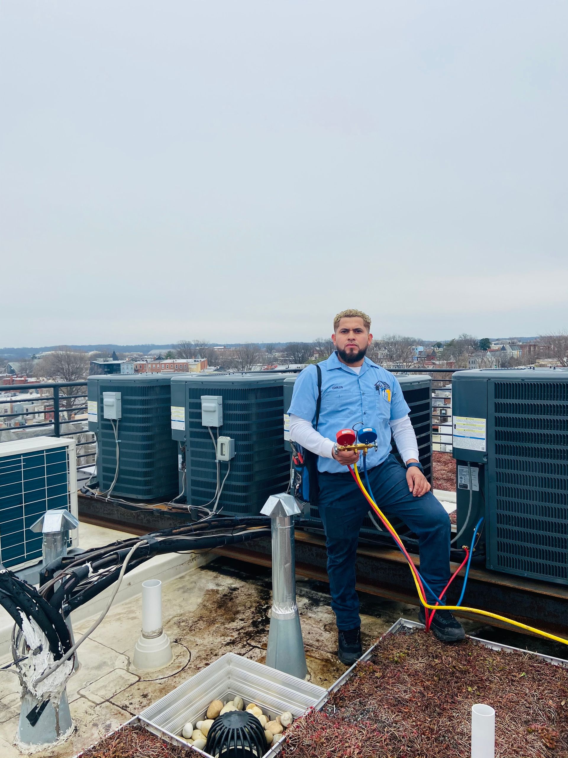 Worker in blue safety gear on a rooftop, holding tools beside HVAC units and pipes.