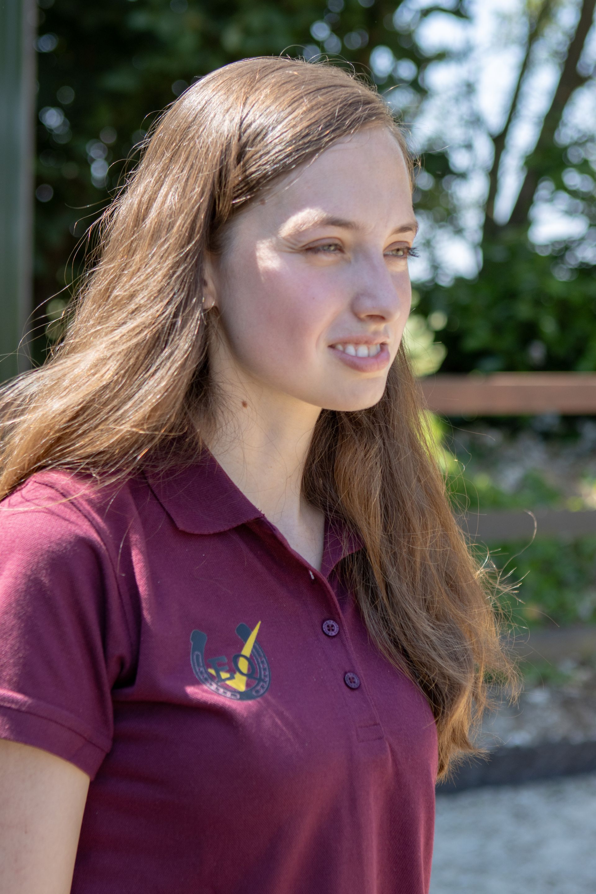 Vrouw met lang bruin haar, gekleed in een kastanjebruin poloshirt, kijkend naar rechts, buiten.