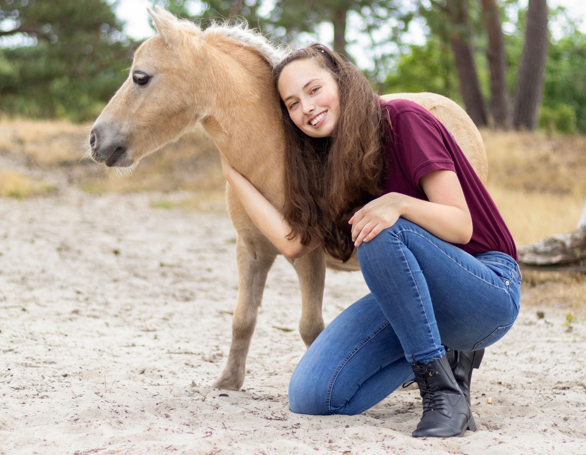 Vrouw omhelst een gebruinde pony op zanderige grond; beiden lachend.